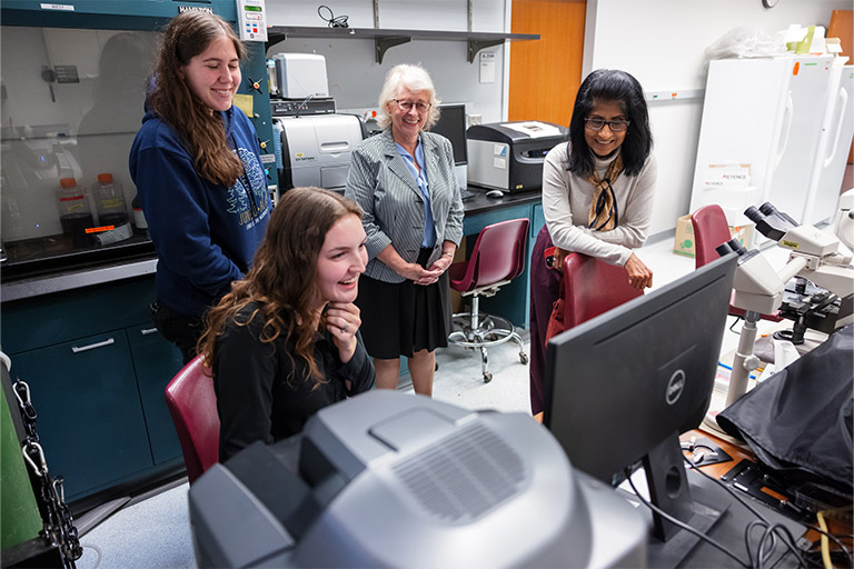 Four people looking at a computer monitor smiling in a laboratory environment.