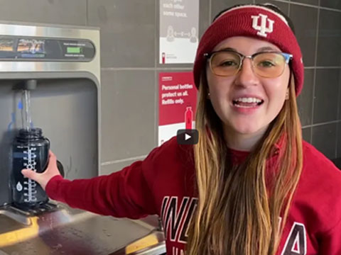 A female student of IUPUI is holding a bottle at water refilling station refilling water and smiling at the camera.